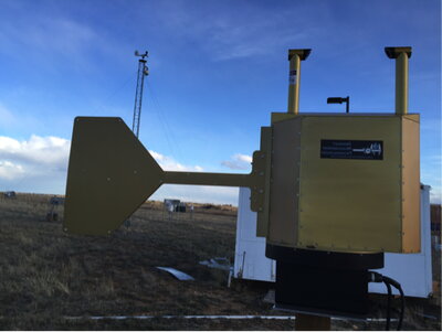 Gold-colored atmospheric monitoring instrument with a directional panel mounted outdoors in an open field.