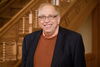 A person in a blazer with a sweater underneath standing in front of a wooden staircase